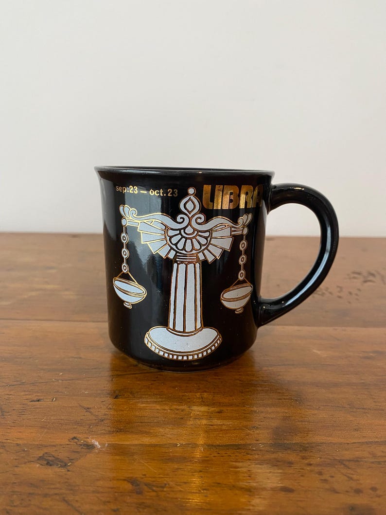 Pode incluir: Caneca de cer&acirc;mica preta com um design do signo do zod&iacute;aco de Libra em dourado e branco. A caneca tem uma al&ccedil;a e a palavra "LIBRA" em letras douradas. As datas "sep.23 - oct.23" tamb&eacute;m est&atilde;o impressas na caneca.