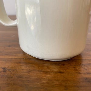 May include: A white ceramic mug with a handle. The mug is sitting on a wooden surface.