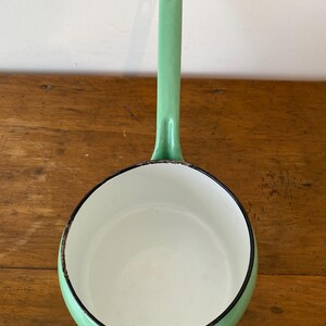 May include: A mint green enamel saucepan with a long handle and a white interior. The rim is outlined in black. The saucepan is sitting on a wooden surface.