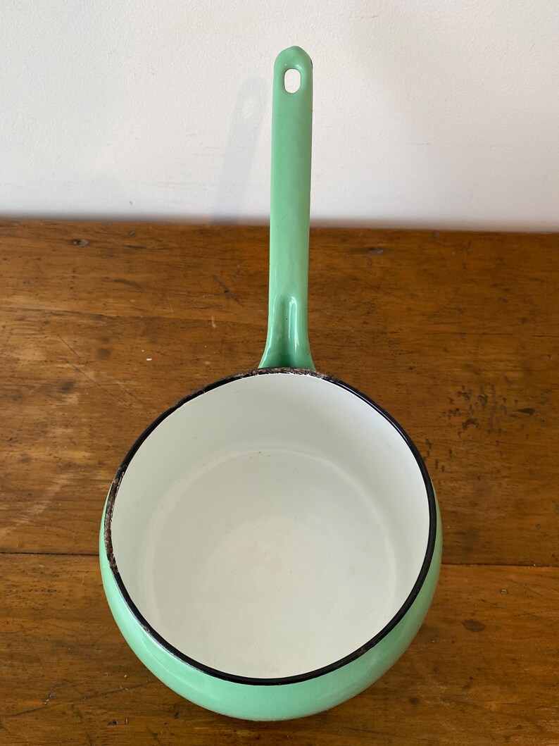 May include: A mint green enamel saucepan with a long handle and a white interior. The rim is outlined in black. The saucepan is sitting on a wooden surface.