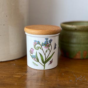 May include: A white ceramic jar with a wooden lid, decorated with a floral and insect design. The illustration features blue and pink flowers, green leaves, and butterflies. The jar is cylindrical and sits on a wooden surface.