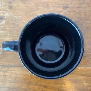 Pode incluir: Uma caneca de cer&acirc;mica preta com acabamento brilhante. A caneca tem um interior arredondado e uma pequena al&ccedil;a. A caneca est&aacute; em uma superf&iacute;cie de madeira.