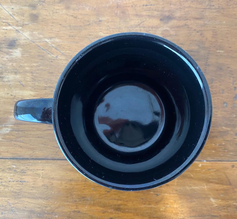 Pode incluir: Uma caneca de cer&acirc;mica preta com acabamento brilhante. A caneca tem um interior arredondado e uma pequena al&ccedil;a. A caneca est&aacute; em uma superf&iacute;cie de madeira.