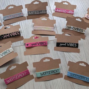 Könnte beinhalten: Verschiedene Haargummis auf Karton-Displays. Die Haargummis sind in verschiedenen Farben und mit Texten wie "good vibes", "limited edition" und "WOTTSEFACK?" in verschiedenen Schriftarten versehen. Der Hintergrund ist eine helle Holzoberfläche.