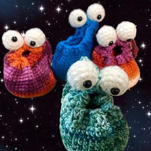 Könnte beinhalten: Fünf farbenfrohe, gehäkelte, außerirdisch aussehende Kreaturen mit großen, weiß-schwarzen Wackelaugen. Die Kreaturen sind in den Farbtönen Lila, Orange, Blau und Grün gehalten. Der Hintergrund ist ein Sternenhimmel.