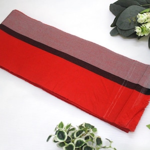 May include: A rectangular red scarf with a gray section and a black stripe. The scarf is made of a soft material and has a fringed edge. The scarf is displayed on a white surface with decorative plants.