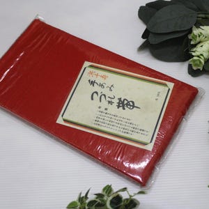 May include: A rectangular, red package wrapped in clear plastic. A label with Japanese text is affixed to the package. Decorative greenery and a patterned ball are in the background.