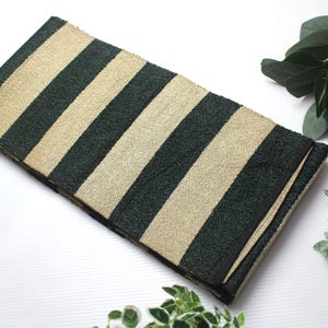 Puede incluir: Un textil rectangular plegado con rayas verde oscuro y beige alternas. El textil está sobre una superficie blanca, con vegetación decorativa y una bola estampada en el fondo.