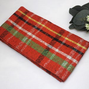 May include: A folded red plaid fabric with white, green, yellow, and black stripes. The fabric is rectangular and appears to be made of woven material. The plaid pattern is evenly spaced and the fabric is neatly folded.