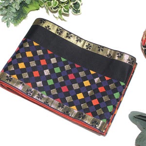 May include: A folded textile featuring a vibrant checkerboard pattern in blue, red, yellow, and green squares, accented with metallic gold squares. The fabric has a wide black border with a gold floral trim along the edges and a red underside.