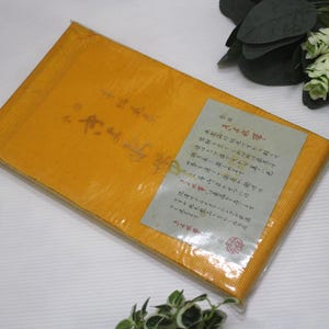 May include: A rectangular, mustard-yellow package wrapped in clear plastic. The package has Japanese text and a label with more text. Decorative greenery and a patterned ball are in the background.