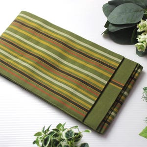 May include: Folded green fabric with horizontal stripes in shades of olive, yellow, orange, and brown. The fabric is folded over, revealing a solid green border. The fabric is likely a decorative textile, such as a table runner or placemat.