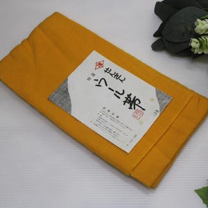 May include: A folded, mustard-yellow textile with a white label featuring Japanese text and a red seal. The textile is rectangular and appears to be made of a textured fabric. Decorative greenery and a patterned ball are in the background.