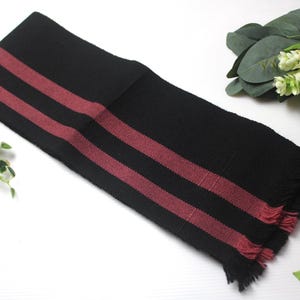 May include: A black scarf with three red stripes, fringed edges, and a rectangular shape. The scarf is displayed on a white surface, with decorative greenery and a patterned ball in the background.