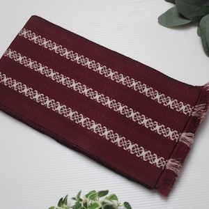 Puede incluir: Textil rectangular burdeos con rayas estampadas blancas. La tela tiene un aspecto texturizado y bordes con flecos. El diseño presenta motivos florales repetidos. El textil es probablemente un artículo decorativo o parte de un proyecto artesanal.