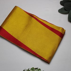 Puede incluir: Una tela dorada doblada con bordes rojos. La tela es rectangular y parece ser de seda o un material similar. Los bordes rojos enmarcan el centro amarillo, creando un contraste llamativo. Es probable que sea un textil decorativo.