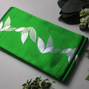 May include: Folded green fabric with a silver leaf pattern. The fabric is a vibrant green color and has a shiny appearance. The leaf design is repeated across the fabric. The image is well-lit and shows the fabric in detail.