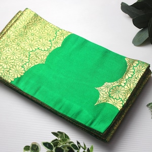 Puede incluir: Un textil verde doblado con un borde dorado con estampado floral. El centro es verde liso y el borde dorado tiene un borde festoneado. El textil es probablemente un artículo decorativo o tela.