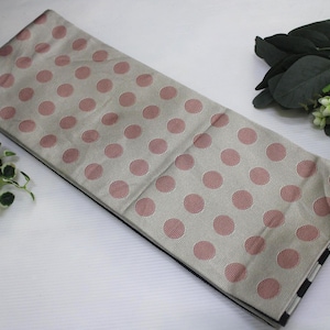 May include: A folded fabric with a beige background and large pink polka dots. The fabric has a black and white striped edge. Decorative greenery and a red and gold ball are in the background.