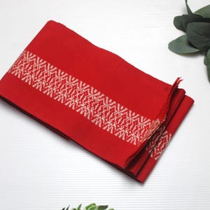 Puede incluir: Un textil rojo doblado con un patrón geométrico blanco. El diseño está centrado en la tela, creando una banda contrastante. El textil es probablemente de material tejido. La imagen está tomada desde una perspectiva cenital.