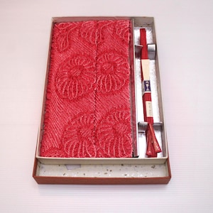 May include: A red textile set in a rectangular box. The set includes a patterned fabric with floral designs and a red decorative cord with a tassel. The box is brown with a white interior.