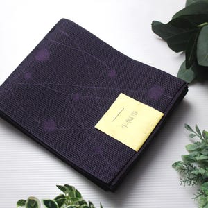 Puede incluir: Un textil plegado, de color púrpura oscuro, con un tejido texturizado y un patrón de círculos y líneas púrpuras. Se adjunta una pequeña etiqueta amarilla con texto japonés. Elementos decorativos: una bola estampada roja y dorada y vegetación.