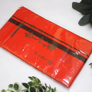 Puede incluir: Un paquete rectangular rojo envuelto en plástico transparente. El paquete tiene una franja negra y texto dorado. El fondo incluye vegetación y una bola decorativa con patrones rojos, negros y dorados.