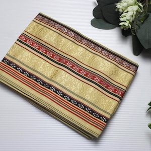 Puede incluir: Tela beige doblada con un estampado floral y detalles a rayas en rojo, negro y blanco. La tela tiene un borde decorativo y probablemente se utiliza para textiles o artesanía.
