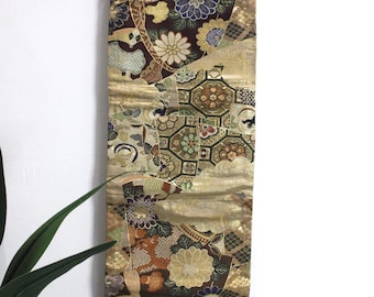 May include: A long, narrow piece of fabric with a gold, brown, and black patterned design. The fabric features a repeating pattern of flowers, geometric shapes, and fans. The fabric is likely a traditional Japanese textile.
