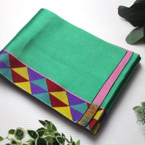 Puede incluir: Un textil verde rectangular doblado con un borde geométrico colorido. El borde presenta triángulos en amarillo, rojo, morado y turquesa. Una franja rosa recorre un borde. El textil es probablemente una bufanda o un paño decorativo.