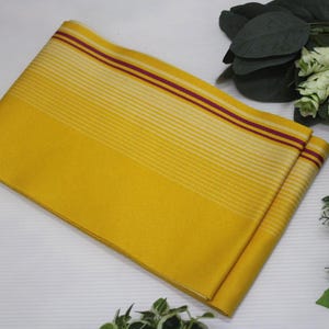 May include: Folded yellow fabric with horizontal stripes and two burgundy stripes near the top edge. The fabric is surrounded by greenery and a decorative ball with red and black geometric patterns.