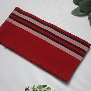 Puede incluir: Un textil rojo doblado con rayas horizontales burdeos y rosa claro. El textil rectangular parece ser de tela tejida. El fondo incluye vegetación y una bola decorativa.