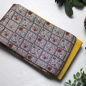 Puede incluir: Un textil plegado con un patrón geométrico plateado y negro, acentuado con motivos florales rojos y una parte inferior amarilla. También hay una bola decorativa roja y dorada y vegetación.