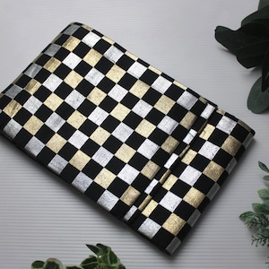 Puede incluir: Un bolso de mano a cuadros negros, plateados y dorados. El bolso de mano es rectangular y está doblado. El patrón es un diseño de tablero de ajedrez repetitivo. El fondo incluye vegetación y una bola decorativa.