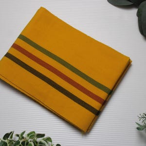 Puede incluir: Tela amarilla doblada con rayas marrones, verdes y rojas. La tela rectangular parece ser tejida. Las rayas corren paralelas a lo ancho de la tela.