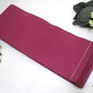 May include: A rectangular, deep magenta fabric with a fringed edge and white stitching. The fabric is folded and lies on a white surface, with decorative greenery and a red and gold ornament in the background.