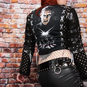 May include: Black leather jacket with silver studs and white lacing, worn with a black mini skirt and high-heeled boots. The jacket's back has a graphic print. Fishnet stockings complete the outfit.