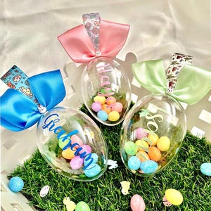 May include: Three clear plastic Easter egg ornaments filled with colourful sweets. Each egg has a different coloured bow and a name written on it. The eggs are on a bed of green faux grass.