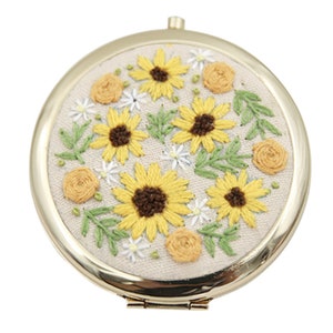 May include: A round, gold-framed mirror with a white fabric background embroidered with yellow sunflowers, green leaves, and white and yellow flowers.