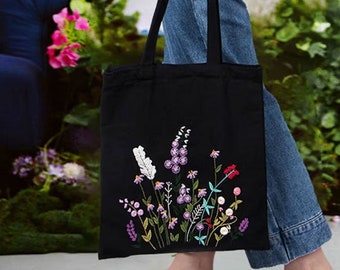 May include: A black canvas tote bag with an embroidered floral design in shades of pink, purple, white, and green. The bag has a long strap and is being held by a person.