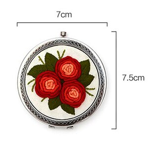 Op de afbeelding: Een ronde borduurring met een witte stoffen achtergrond en een bloemenontwerp van drie rode rozen met groene bladeren. De ring heeft een diameter van 7 cm.