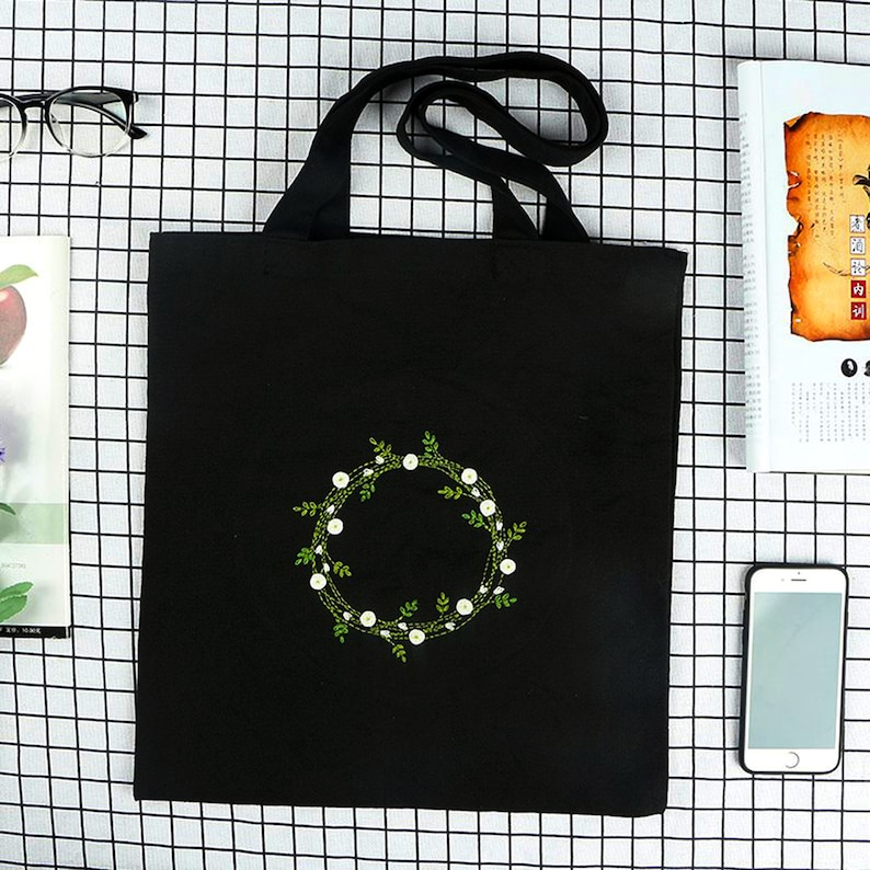 May include: Black canvas tote bag with a white floral wreath embroidered on the front. The wreath is made of small white flowers and green leaves.