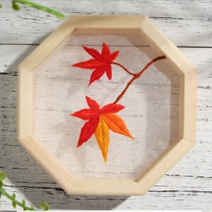 Peut inclure: Feuilles d'automne brod&eacute;es dans des tons d'orange et de rouge sur un fond blanc, encadr&eacute;es dans un octogone en bois naturel.