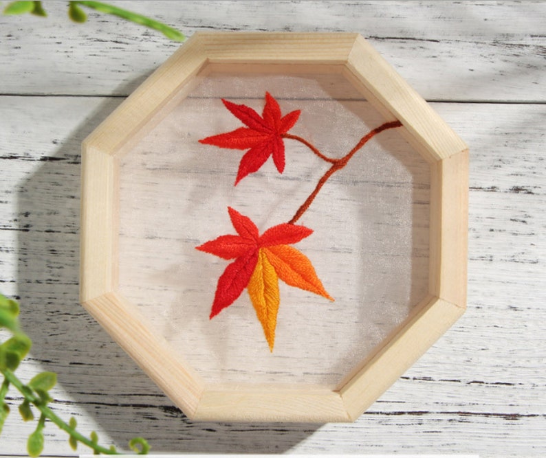 Peut inclure: Feuilles d'automne brod&eacute;es dans des tons d'orange et de rouge sur un fond blanc, encadr&eacute;es dans un octogone en bois naturel.