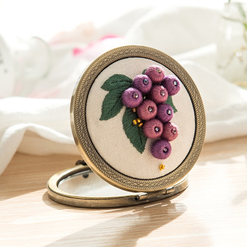 Op de afbeelding: Een kleine, ronde, antieke goudkleurige compactspiegel met een wit stoffen interieur met een geborduurd ontwerp van een tros paarse druiven met groene bladeren.