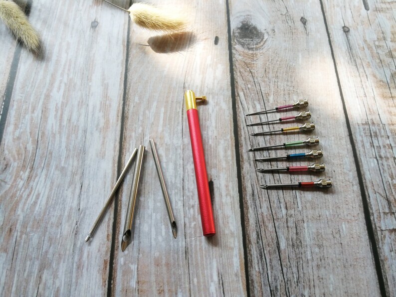 May include: A set of 10 embroidery needles with colorful plastic handles and a red and gold handled needle tool. The needles are arranged on a wooden surface.