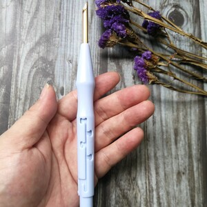 Op de afbeelding: Een close-up van een lichtpaarse plastic garennaald met een gouden metalen punt. De naald wordt in een hand gehouden tegen een houten achtergrond.