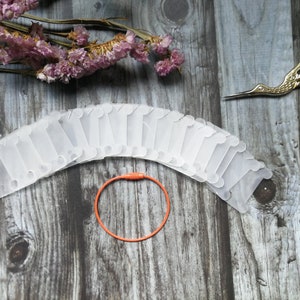 May include: A set of 20 white plastic thread bobbins arranged in a semi-circle on a wooden surface. A single orange rubber band is in the centre of the bobbins.