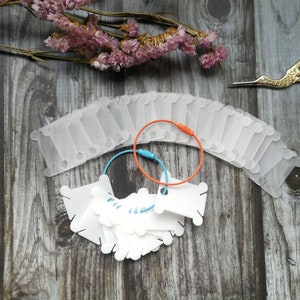 Könnte beinhalten: Ein Satz weißer Plastik-Garnrollen mit einem blauen und orangefarbenen Band. Die Rollen sind halbkreisförmig auf einer Holzoberfläche angeordnet.