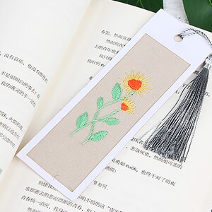 May include: A beige bookmark with a white border and a tassel. The bookmark features an embroidered design of a green stem with two yellow flowers.